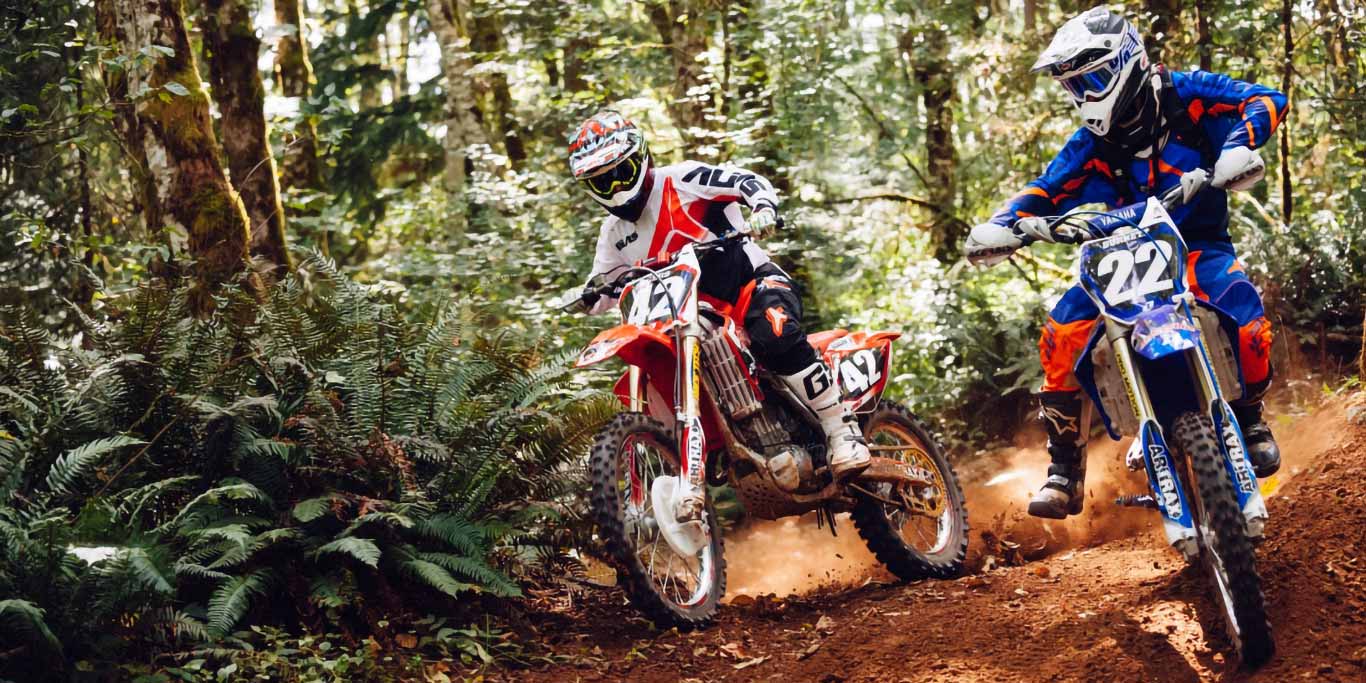 Two dirt-bike riders race through a forrest setting.
